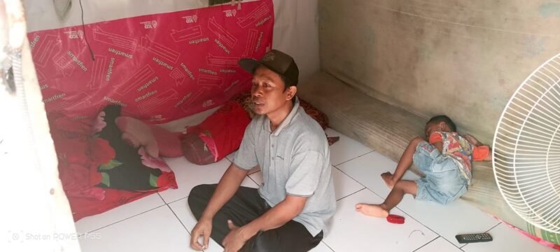 Arif duduk di dalam ruangan 3x3 meter berdinding bilik bambu yang ditinggalinya bersama istri dan dua anak. Rumah sempit ini menjadi tempat mereka bertahan hidup di tengah keterbatasan ekonomi